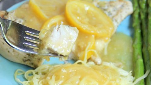 A plate of garlic lemon chicken garnished with lemon slices, served with pasta in a yellow sauce and asparagus. A fork holds a bite of chicken above the plate. Text on the image reads “Garlic Lemon Chicken.”.